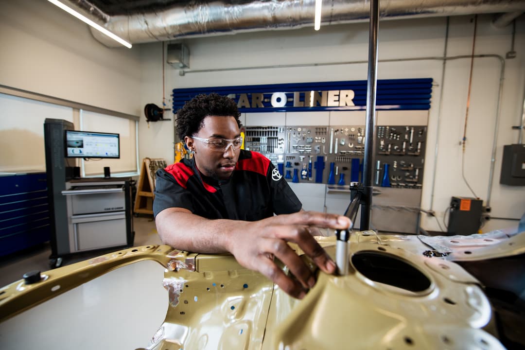 Auto body student in safety glasses works on a car frame in a Car-O-Liner training shop with tools on the wall.