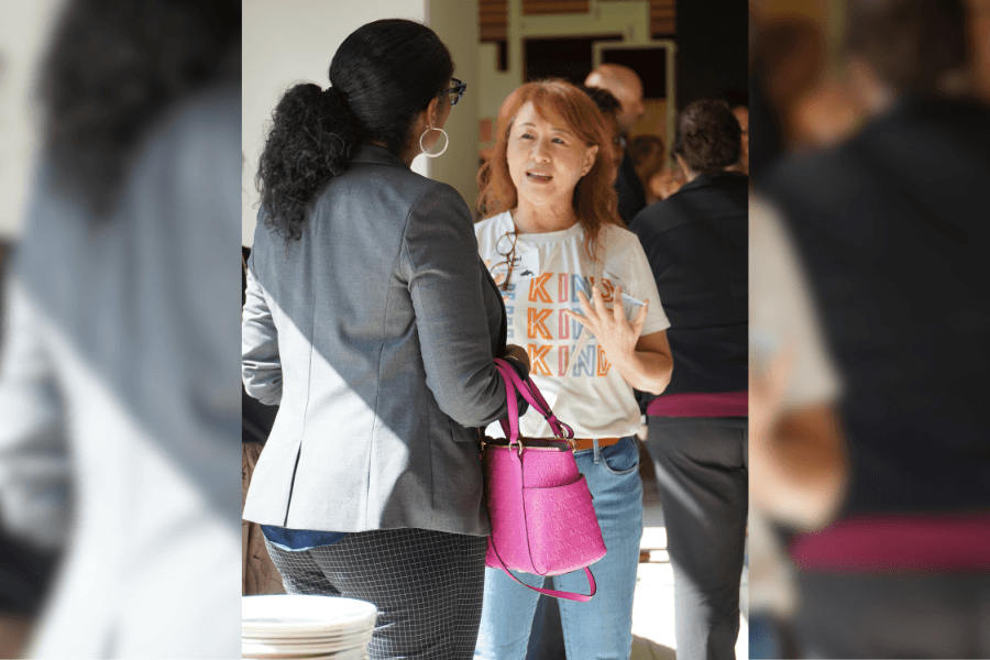 Two women talk at a crowded gathering, one in a gray blazer with a pink handbag, the other in a "Kind" t-shirt.