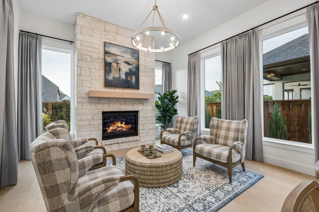 Living room with plaid armchairs, stone fireplace, round woven ottoman, and large windows with gray curtains.