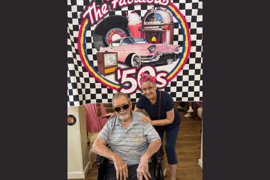 Man in wheelchair with sunglasses and woman in red headband pose by a "Fabulous '50s" backdrop.