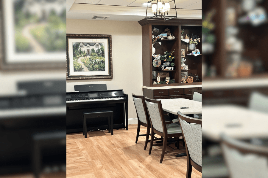 Common room with digital piano, framed cottage painting, dining table with chairs, and wooden display cabinet.