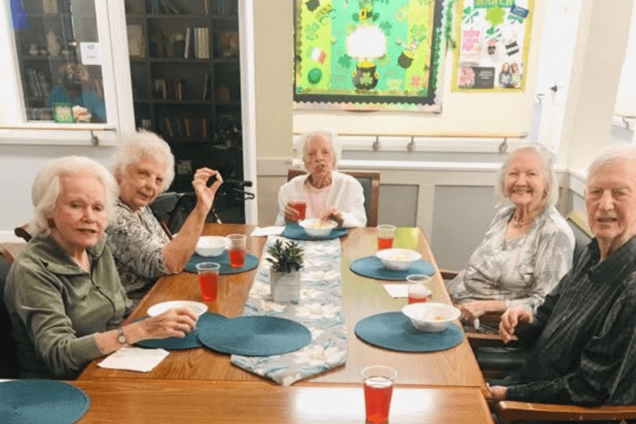 Five elderly people seated at a table with bowls and drinks, near a St. Patrick's Day bulletin board.