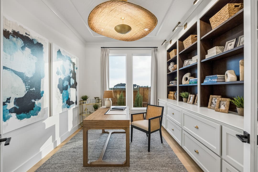 Home office with wooden desk, cane chair, rattan pendant light, built-in shelves, and blue abstract wall art.