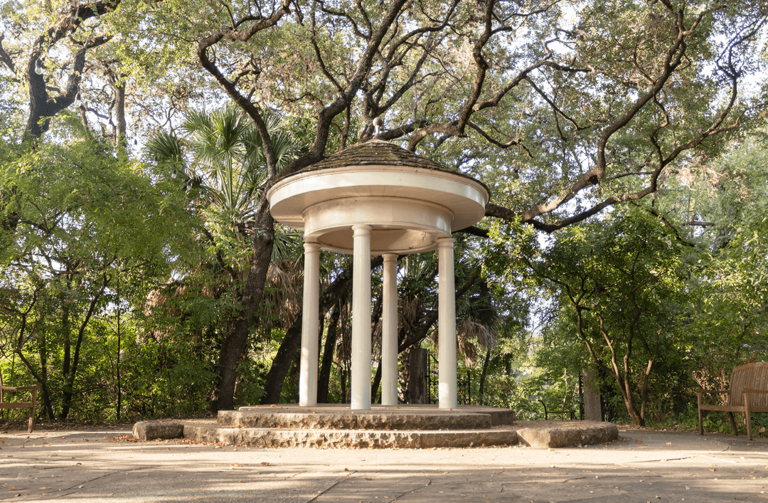 White circular gazebo with columns on stone steps, surrounded by lush green trees in a park.