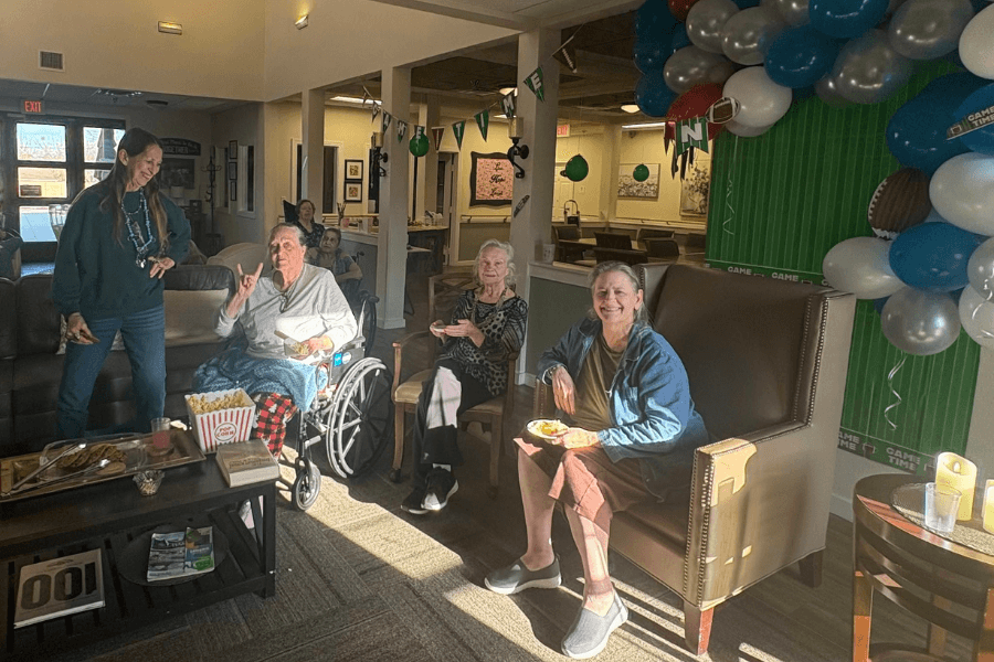 Seniors gather for a football-themed party with balloons, popcorn, and snacks in a care facility common room.