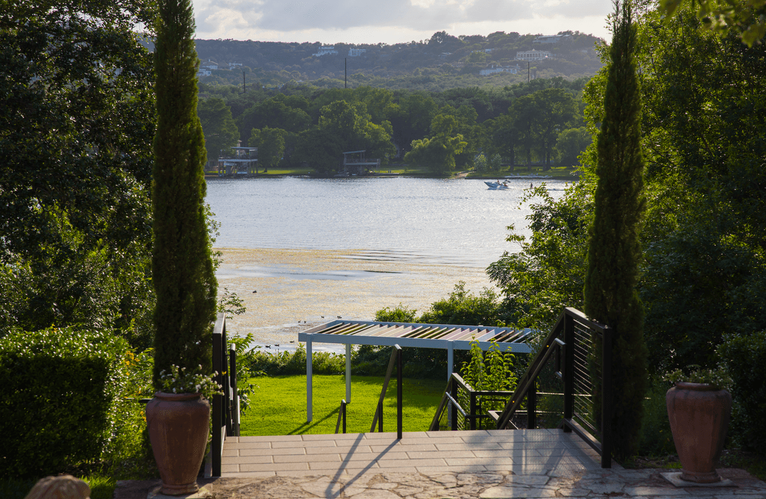 Lake view framed by cypress trees and terracotta urns, with a pergola, boat on water, and hills beyond.