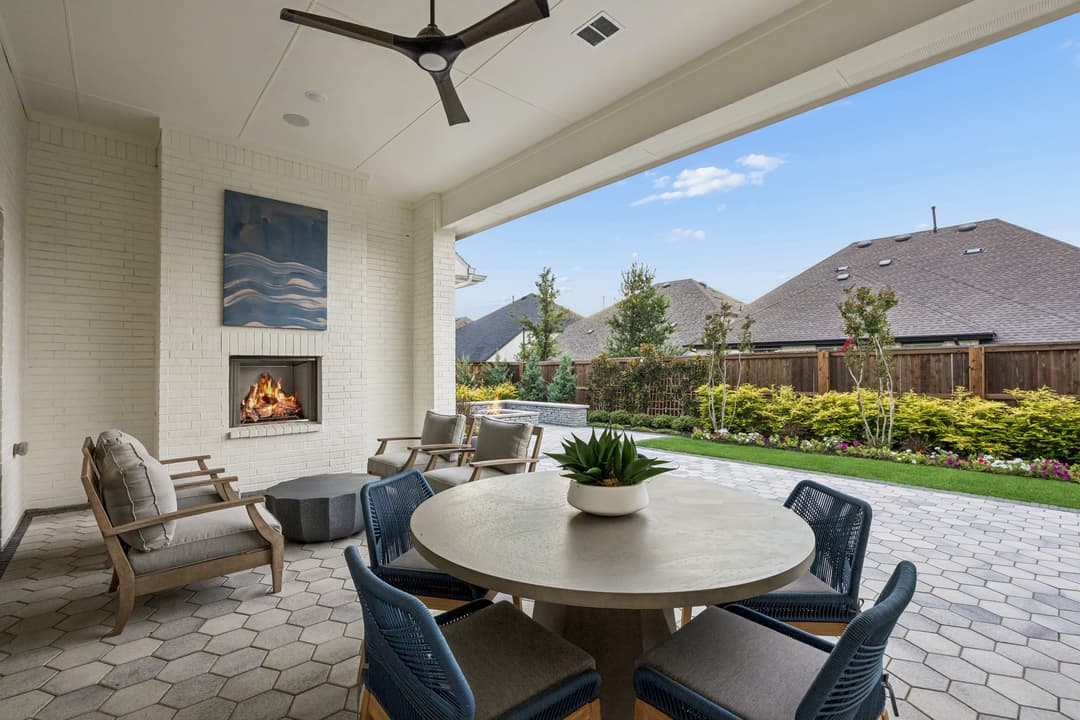 Covered patio with dining table, chairs, lit brick fireplace, and ceiling fan overlooking a landscaped backyard.