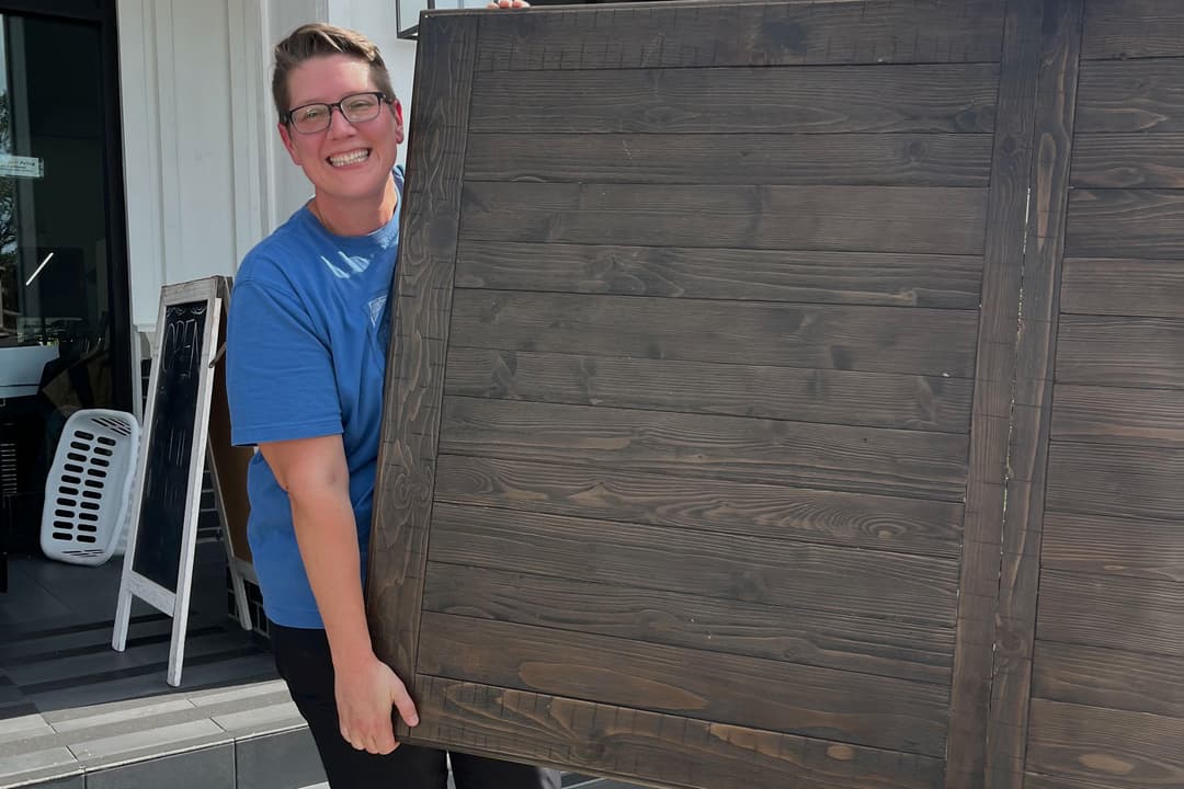 Smiling person in blue shirt and yellow shoes carries a large wooden tabletop down steps outside a shop.