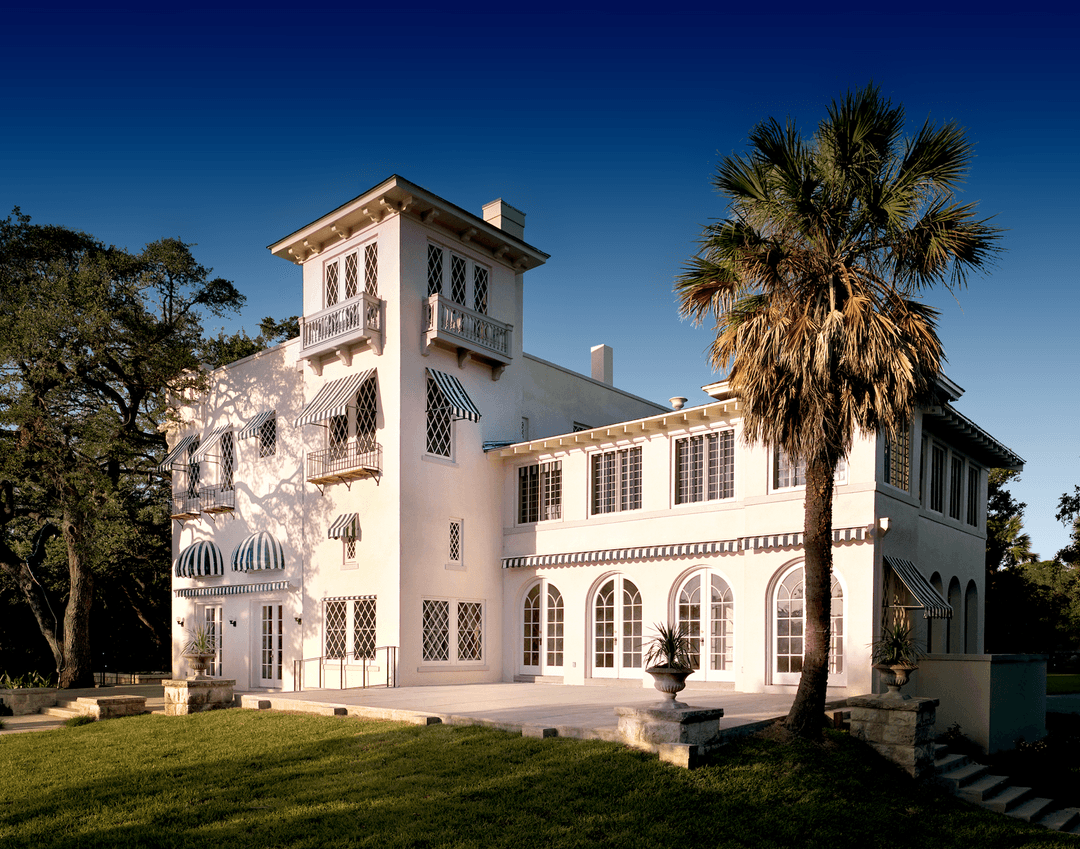 Mediterranean-style mansion with tower, striped awnings, and arched windows beside a palm tree under clear blue sky.