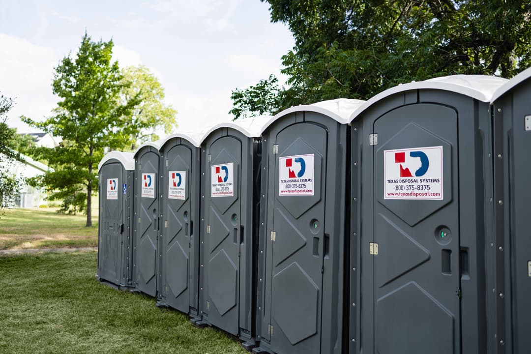 A row of gray Texas Disposal Systems portable toilets lined up on grass beneath trees.