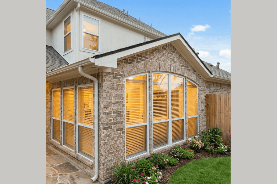 Brick two-story home exterior with large arched windows, warm interior lighting, landscaped flower beds, and a stone patio.
