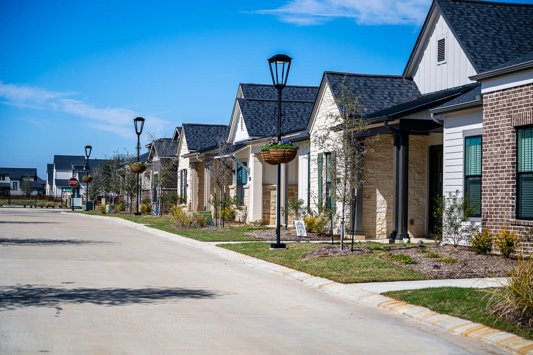 Row of new stone and brick homes along a quiet street with lampposts and hanging flower baskets under a blue sky.