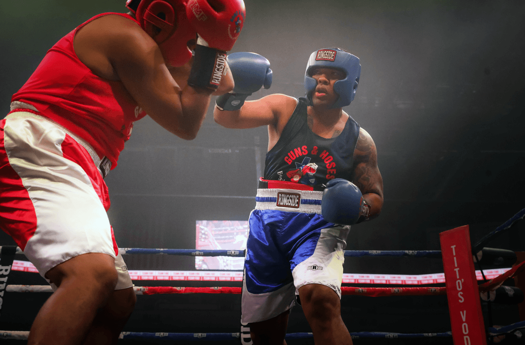 Boxer in blue "Guns & Hoses" gear throws a jab at an opponent in red who blocks with gloves up in the ring.