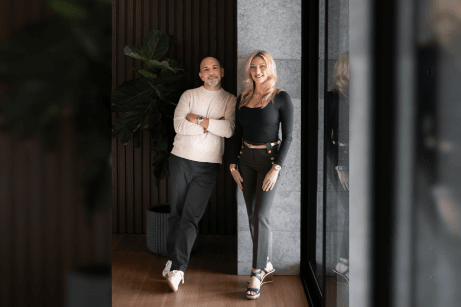 A man and woman in stylish attire pose together indoors near a large plant and modern wall.