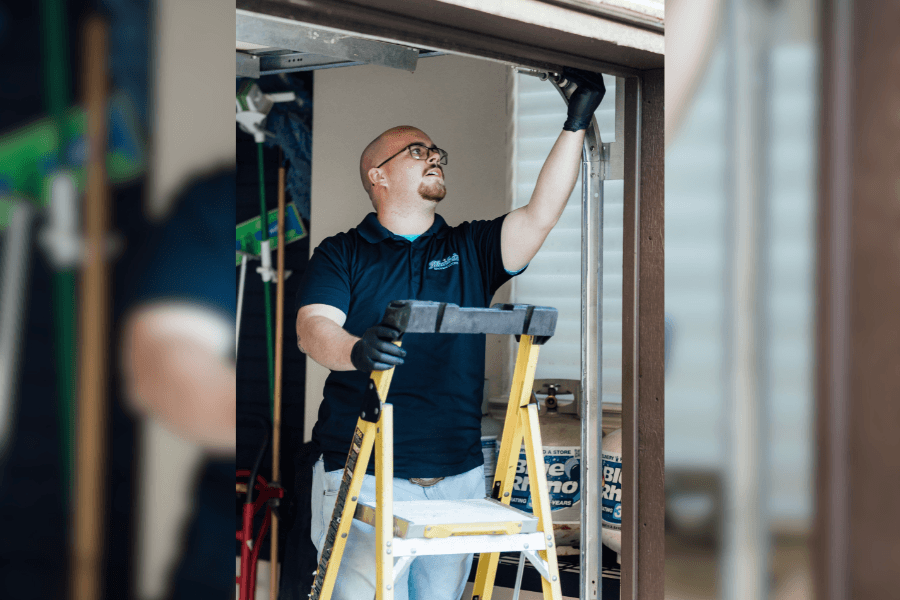 Technician in gloves repairs an overhead garage door track while standing at a ladder in a garage.