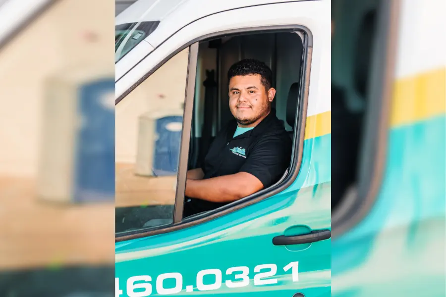 Smiling man in black work shirt sits in the driver's seat of a teal and white company van.