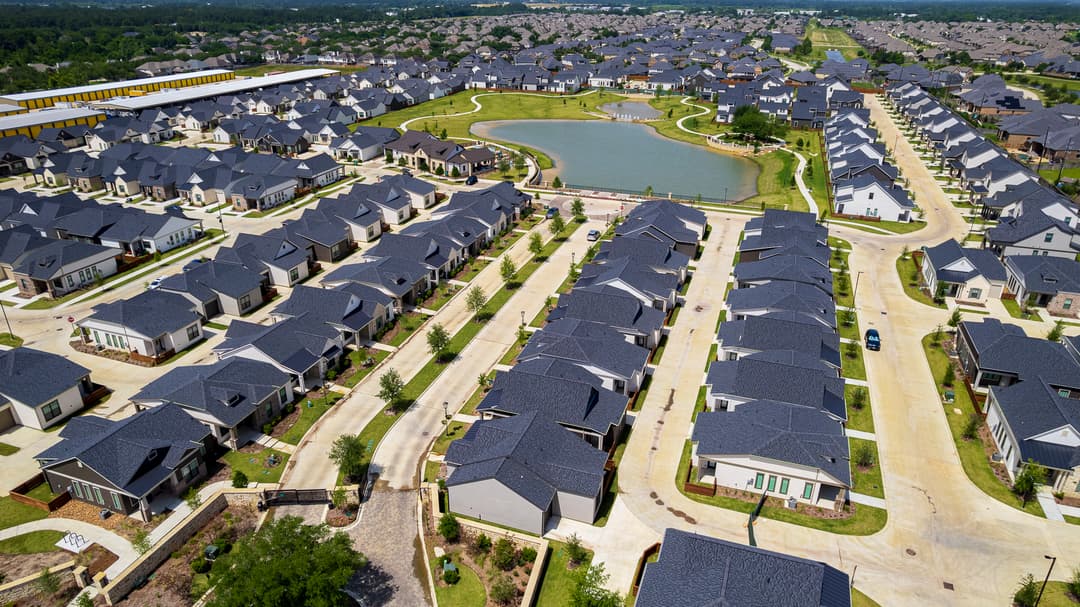 Aerial view of a suburban housing development with rows of gray-roofed homes surrounding a small pond.