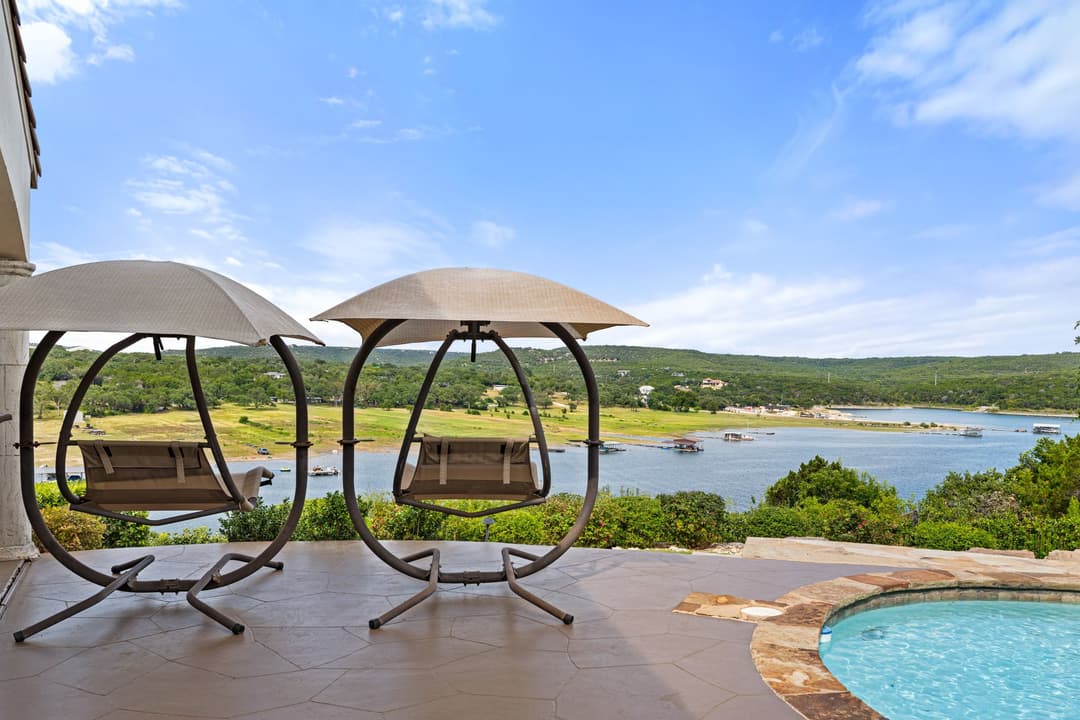 Two canopied swing chairs on a stone patio beside a pool overlooking a lake and green hills.
