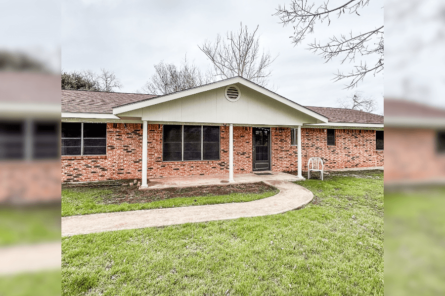 Single-story brick house with covered front porch, curved walkway, and grassy lawn under an overcast sky.