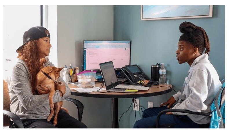 Two women talk at an office table with a laptop and monitor; one holds a small dog on her lap.