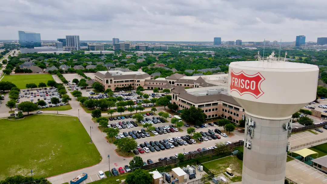 Aerial view of Frisco, Texas water tower overlooking office buildings, parking lots and distant city skyline.