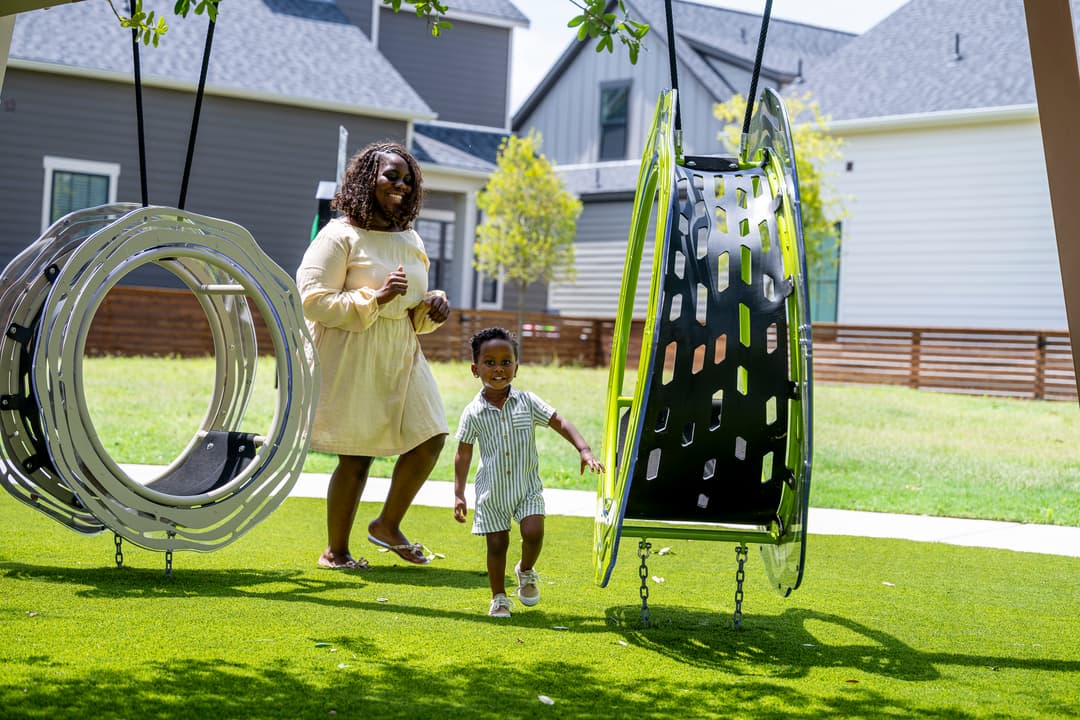 Smiling child runs between modern playground swings as a woman follows on a sunny grassy lawn.
