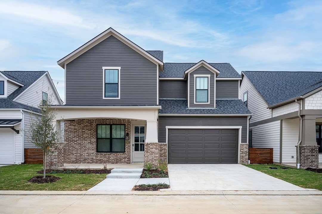 Two-story gray home with brick porch, two-car garage, and concrete driveway in a new residential neighborhood.
