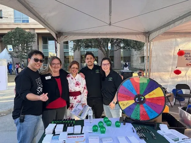 Five people pose behind an event booth with a colorful prize wheel, flyers, and giveaways under a tent.