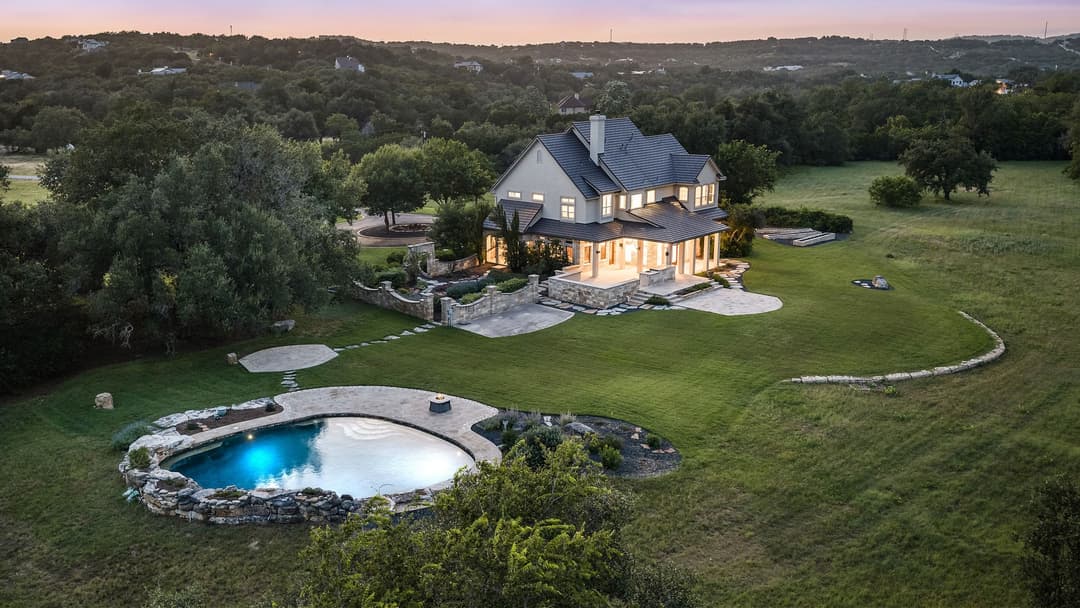 Aerial view of large two-story house with lit windows, stone-edged pool, and green lawn amid wooded hills at dusk.