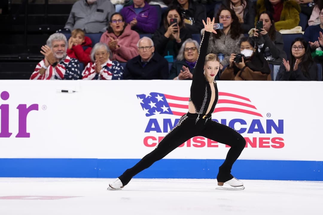 Figure skater in a black costume strikes a pose with arm raised as spectators applaud behind the rink boards.