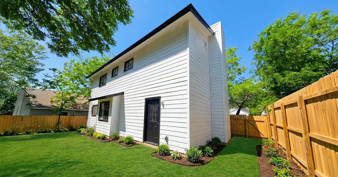 Two-story white house with black door in a fenced backyard with green lawn and landscaped flower beds.