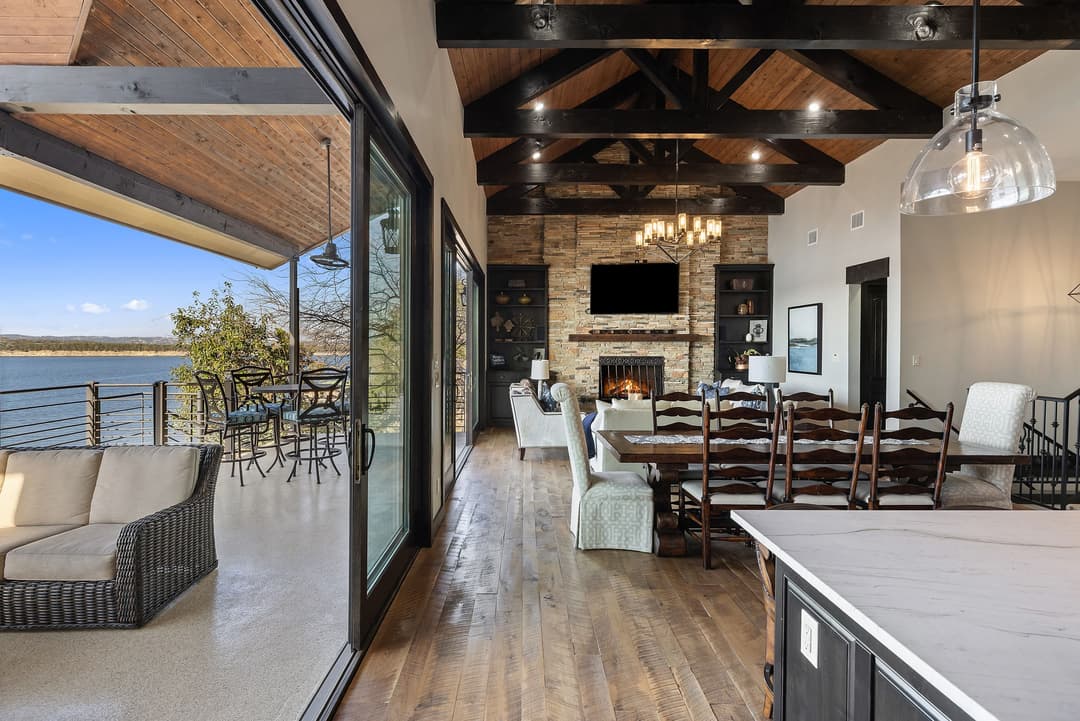 Open living and dining area with vaulted wood-beam ceiling, stone fireplace, and sliding doors to a lakeside patio.
