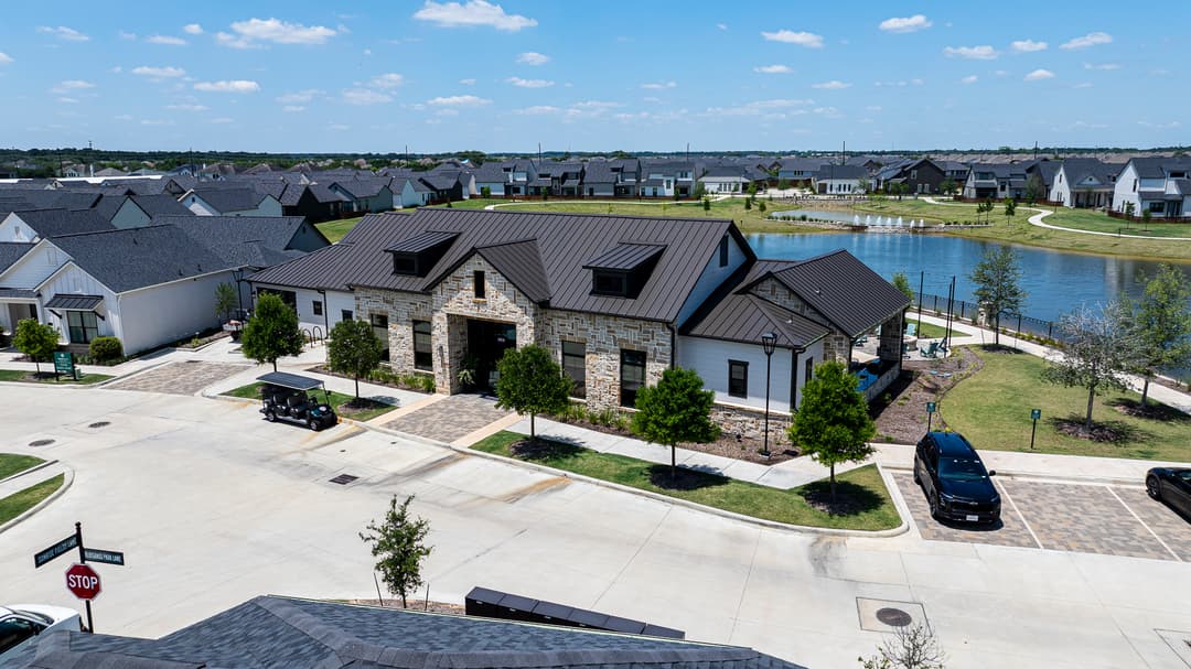 Aerial view of a stone community clubhouse beside a lake in a suburban neighborhood, with golf cart and parked cars.