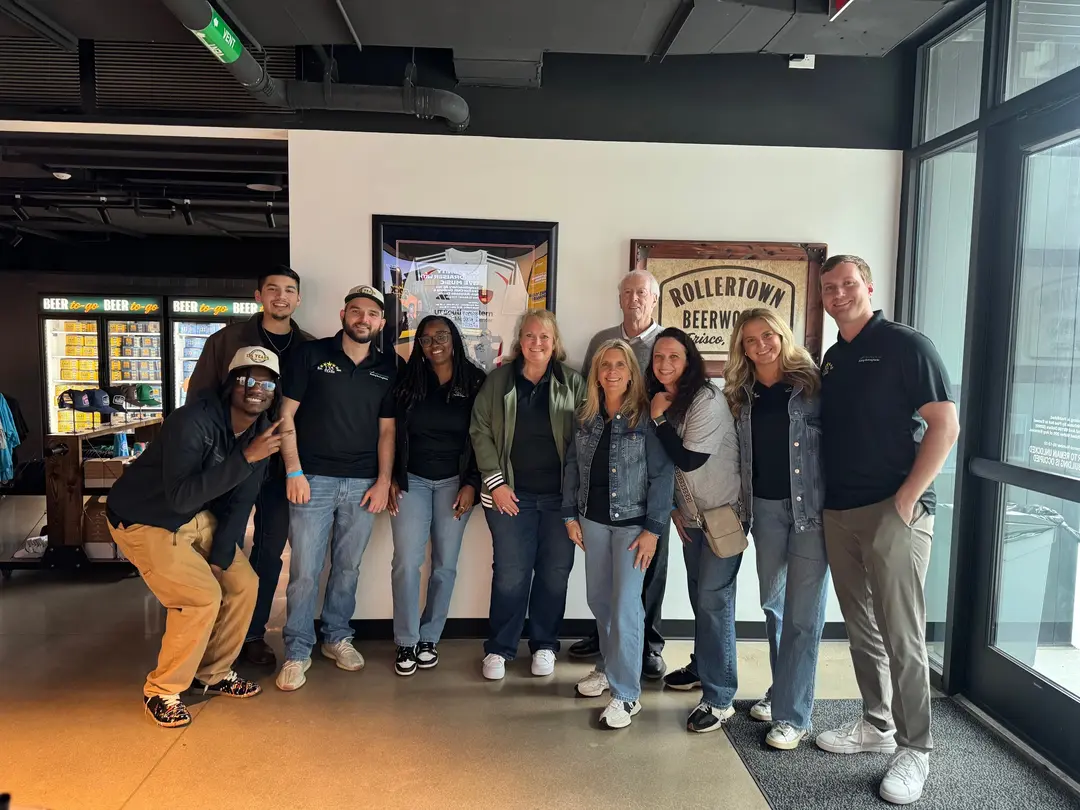 Group of eleven people smiling together in front of a Rollertown Beerworks sign at a Frisco brewery.