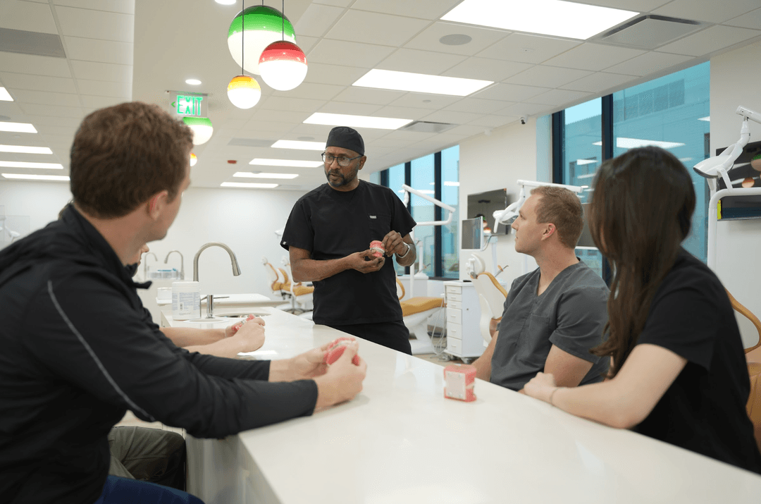 Dentist in scrubs holds a dental model, teaching three students seated at a counter in a modern dental clinic.