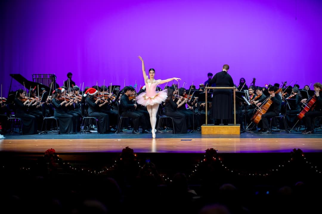 Ballerina in pink tutu on pointe performs before an orchestra and conductor at a holiday concert.