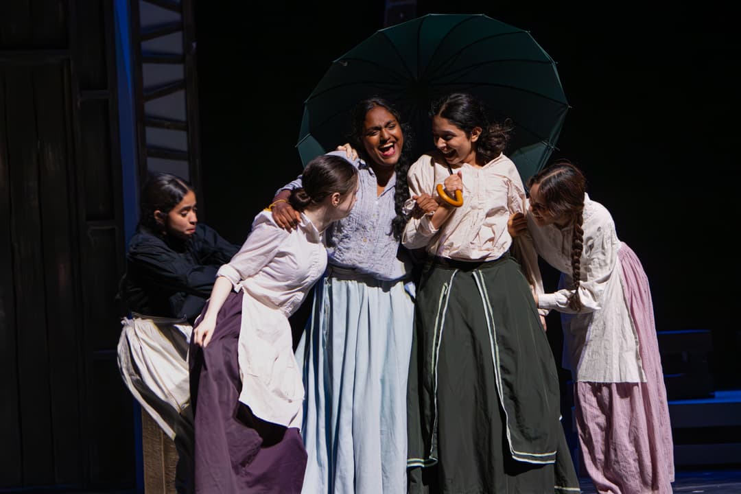 Actors in period costumes laugh together under a green umbrella on stage, while one performer sits apart in the background.