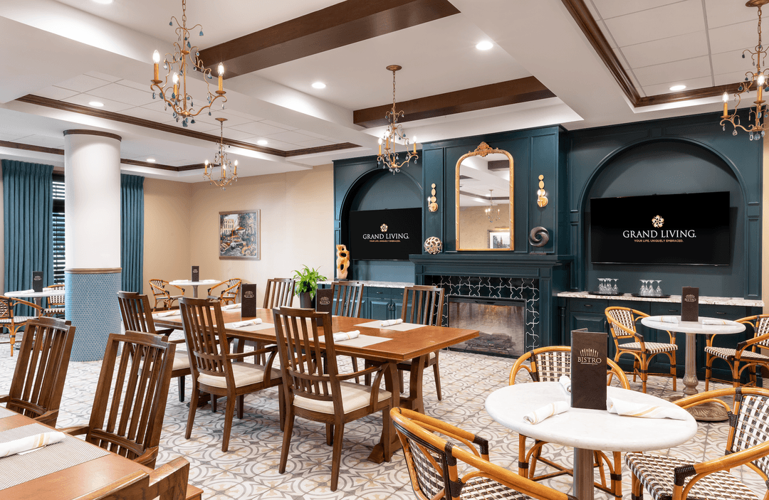Upscale bistro dining room with wooden tables, chandeliers, fireplace, mirror, and Grand Living signs on TV screens.