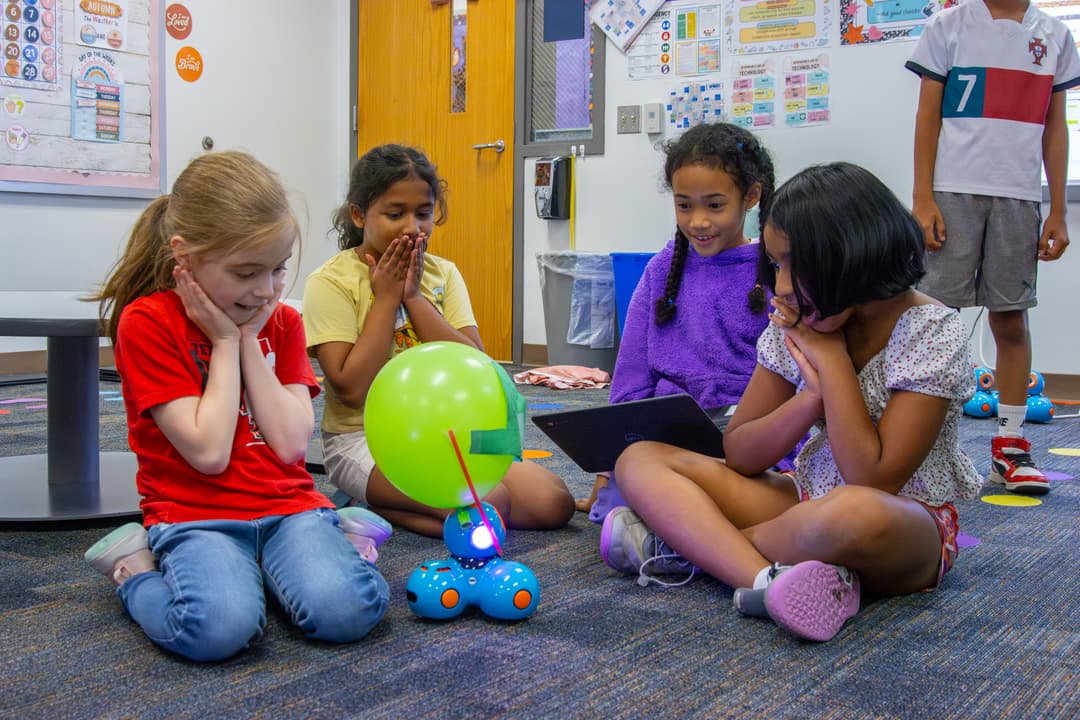 Four children sit on classroom floor watching a blue robot with a green balloon attached, using a laptop.
