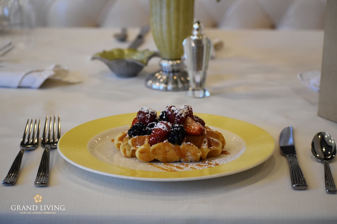 Waffle topped with mixed berries and powdered sugar on a yellow plate with silverware on a linen table.