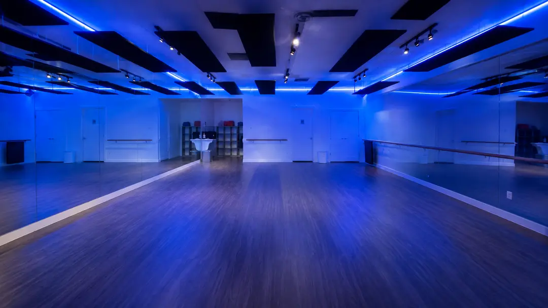 Empty dance studio with wood floor, mirrored walls, ballet barres, and blue LED lighting on the ceiling.