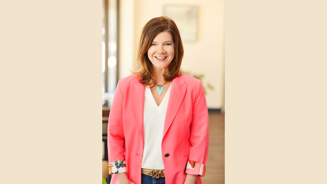 Smiling woman with brown hair wearing a pink blazer, white top, and turquoise heart necklace indoors.