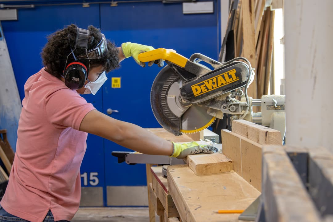 Person in mask, goggles, ear protection and gloves cutting wood on a DeWalt miter saw in a workshop.