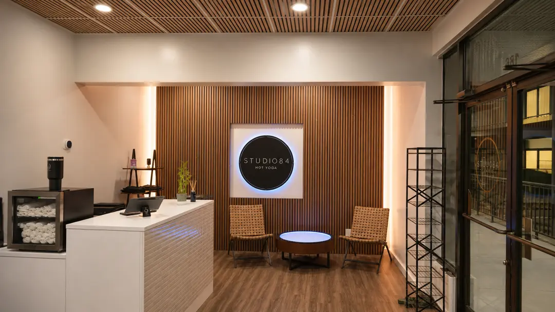 Studio 84 Hot Yoga lobby with reception desk, wooden slat wall, illuminated sign, and two wicker chairs.