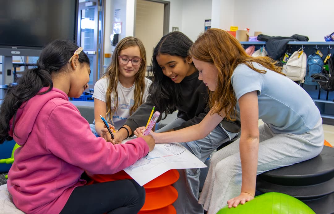 Four girls gather around and work together on a drawing with pencils in a classroom setting.