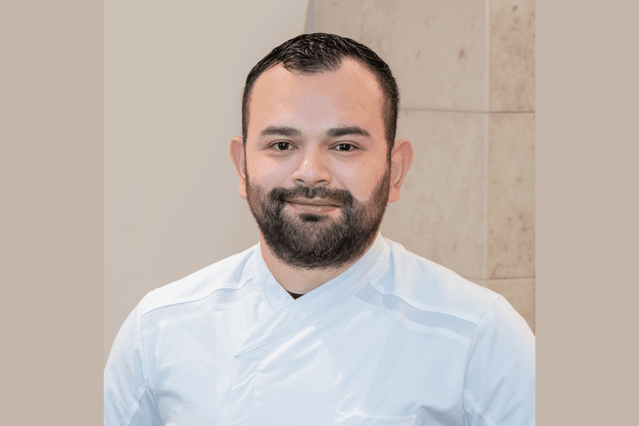 Man with beard wearing a white chef's or medical jacket, smiling in a professional headshot.