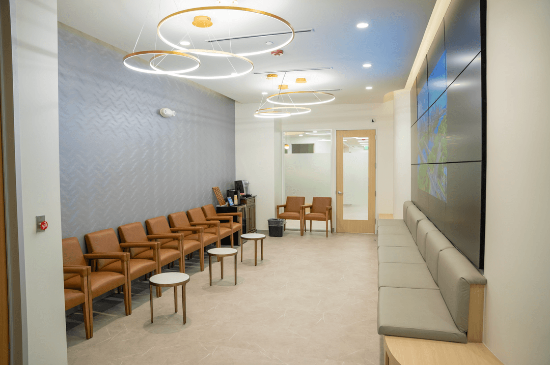 Modern waiting room with tan leather chairs, gray bench, ring lights, wall screen and glass door.