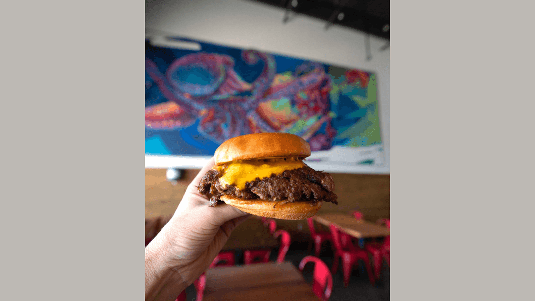 Hand holding a cheeseburger inside a restaurant with a colorful octopus mural and red chairs.