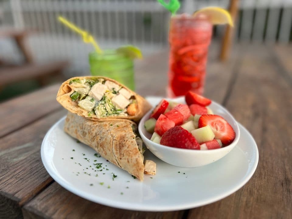 Chicken Caesar wrap with a side of fresh fruit and two drinks on an outdoor wooden table.