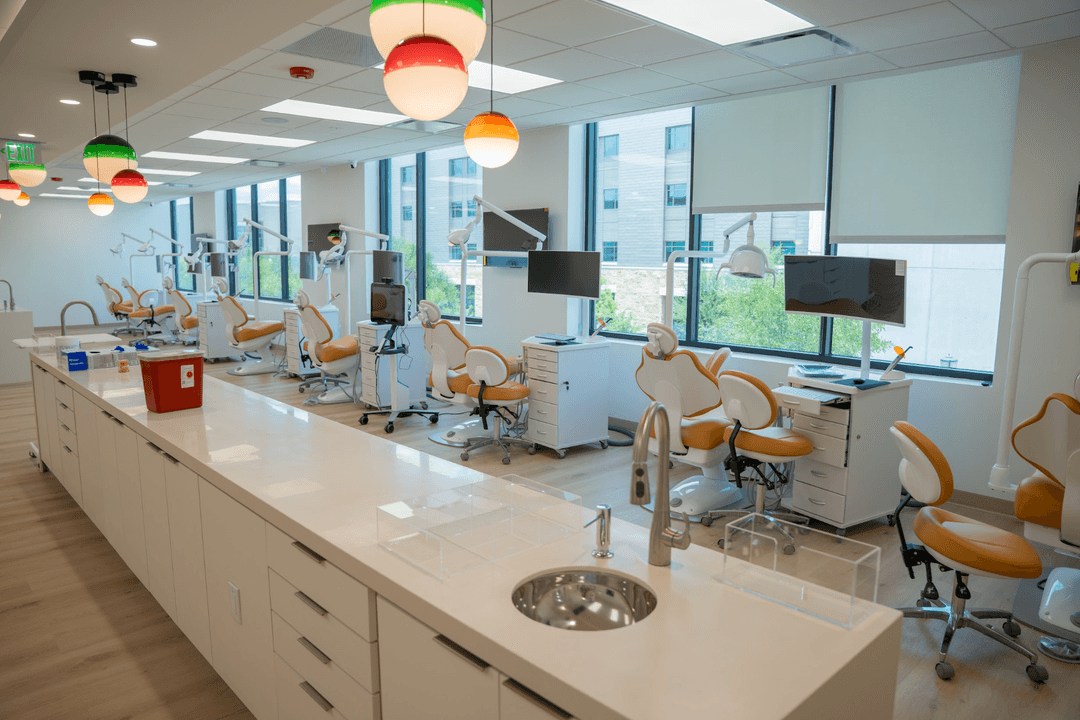 Modern dental clinic with rows of tan patient chairs, exam lights, monitors, and colorful pendant lamps by windows.
